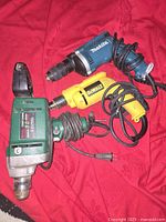 Three electric corded drills placed on red cloth, brands Makita (blue), DeWalt (yellow), Century (green metal) visible. Cords coiled beside each.