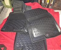 Four black rubber car floor mats laid on a red cloth surface showing textured pattern and edges, including front mats with retention clip area.