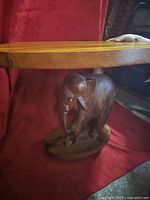 Side view of wooden elephant carving supporting a wooden tabletop against a red fabric background.