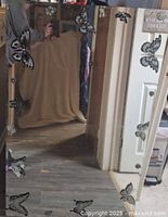 Photo of a tall rectangular mirror showing butterfly decals around the edges. The mirror reflects part of a room including floor and door.