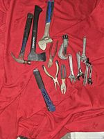 Photo showing mixed hand tools arranged on a red fabric background including hammers, adjustable wrenches, pliers, a puller tool, and a folding ruler.