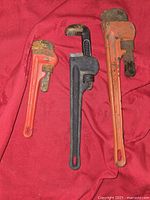 Three pipe wrenches laid out on a red cloth background. Small red wrench on left, medium black wrench in center, large red/orange wrench on right. All show various degrees of rust and wear on jaws.