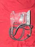 Photo of red electric handheld sander with rectangular sanding pad and connected power cord, placed on red fabric background.