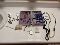 Full view of two trays containing various bracelets, necklaces including long strands of black and cream beads, earrings, and a white pearl-like stretch bracelet.