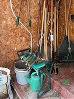 Photo showing various wood-handled garden tools including rakes, hoes, axes, and shovels leaning against a wooden shed wall, along with a green watering can, buckets, and a small garden bench with hand tools resting on it.