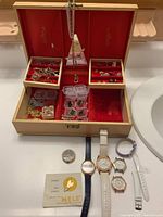 Photo of open jewel box with red velvet interior full of assorted costume jewelry including earrings, chains, pendants, rings, and small plastic boxes with jewelry parts. Five untested wristwatches and a watch booklet are displayed in front of the box.