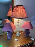 Three lamps displayed on a wooden surface with mirror behind; one tall clear plastic and glass based lamp in center with burgundy shade and two smaller burgundy bedroom lamps with matching fabric shades on either side.