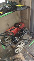 Photo showing the entire lawnmower with two red jerry cans next to it, placed on the shed floor with some garden tools overhead.
