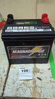 Close-up of one black Magnacharge lawn mower battery showing positive red and negative black terminals and brand label.