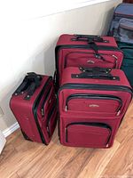 Three red fabric suitcases in a row leaning against a white wall on wooden floor.