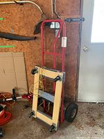 Two wheeled red metal hand truck dolly with wooden platform and large black rubber wheels leaning against wall