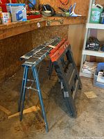Four saw horses - two metal blue and two plastic black with orange tops, used condition with paint and wear marks, placed in a garage with shelves and materials.