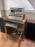 Stereo setup with Koss DVD player, Sanyo cassette deck and receiver stacked on top of wood media cabinet, Sanyo turntable visible inside cabinet, two floor model speakers upright on either side of cabinet, cables inside cabinet.