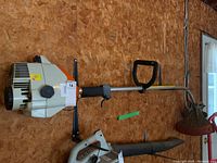 Side view showing full length gas powered trimmer with curved shaft, handle grip, and cutting head.