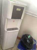 Front view of the white Hamilton Beach water dispenser showing the gray control panel with buttons and dispensing area with drip tray.