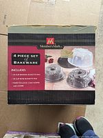 Box of Members Mark 4-piece bakeware set showing contents: two 10 cup bundt pans (Bavaria and Rose styles) and twist-to-lock cake keeper.