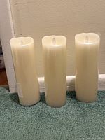 Photo showing three ivory-colored cylindrical battery operated candles with LED flickering flame tops placed on a green carpet surface against a beige wall.