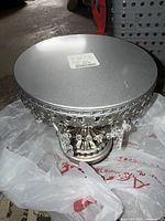 Top-down and angled view of silver metal decorative cake stand with scalloped edge and hanging clear crystal pendants on a white plastic bag.