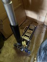 Blue and silver hydraulic floor jack with handle and pressure gauge, placed on a concrete floor near cardboard.