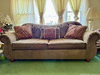 Front view of Raymour & Flanigan sofa with three paisley patterned throw pillows, showing textured grey upholstery, wooden bun feet, and button tufting on the backrest.