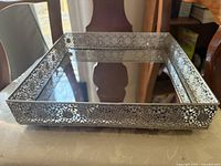 Rectangular mirror tray with ornate silver filigree metal border, reflective mirrored base, sitting on a table.