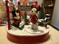 Close-up of Disney Christmas decoration featuring Mickey Mouse on a sleigh and Minnie skating, with snow-covered base and pine trees.