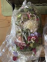 Front view of the white metal birdcage with colorful silk flowers inside, covered with plastic wrap.