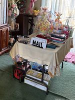 Photo of a small folding table covered with a beige tablecloth and various items on top, including mail and wrapped gifts. The table has a black metal frame and appears stable.