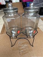 Photo of two clear hexagonal glass drink pitchers with lids and silver spigot taps, sitting on black metal stand. Several small plastic cups are wrapped in plastic and placed in front of stand.