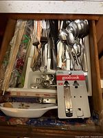 Overhead photo of a kitchen drawer containing various kitchen utensils including forks, spoons, knives, chopsticks, bottle openers, and a ceramic dish