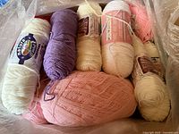 Photo showing multiple skeins of yarn in white, purple, beige, pink, and peach colors in a large box, wrapped in plastic.