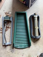 Photo of two lawn sprinklers, a green downspout diverter, and a gray flexible gutter extension hose laid out on a concrete surface with leaves in the background.