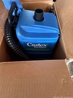 Photo of blue Castex Portapac backpack vacuum cleaner inside a cardboard box, includes coiled black hose and ergonomic handle for carrying.