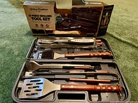 Opened carrying case showing all 17 grilling tools neatly arranged inside, including spatula, fork, tongs, cleaning brush with wooden handles and stainless steel construction.