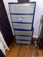 Plastic Sterilite drawer cart with multiple translucent drawers and dark blue framing on wooden floor next to a wall with electric cords nearby.