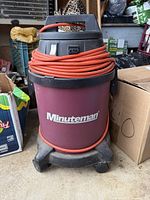 Front view of maroon Minuteman shop vacuum with black top and orange extension cord wrapped at the top.