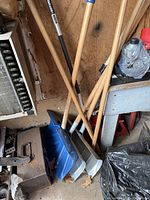 Photo showing five shovels with wooden handles leaning against a wall, including two snow shovels with plastic blades.