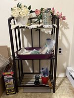 Full view of the small dark metal shelf showing all four shelves with contents including decorative flowers, glass jar with beads, plastic bags with beads, hose attachment, and small parts scattered.