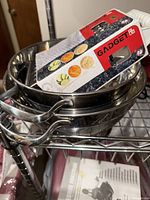 Photo shows two stainless steel strainers stacked together with handles visible, and a handheld stainless steel grater in original packaging on top.