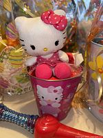 Hello Kitty plush toy seated in a pink metal bucket with Hello Kitty design, surrounded by decorative yellow ribbons.