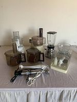 Photo showing assorted small kitchen appliances on a table including multiple food processors, a blender, and hand blenders.