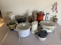 Full view of all kitchen appliances on table including slow cookers, crockpot, kettle, food processor, coffee maker, and hand blender