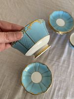 Hand holding one teacup showing powder blue panels with gold scrollwork, two saucers visible on table
