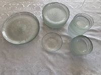 Top-down view of 5 dinner plates, 8 side plates, and 15 small bowls arranged on a white patterned tablecloth showing clear glass material with floral textured design and scalloped edges.