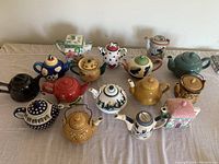 Full collection of 14 colorful ceramic teapots arranged on a cloth-covered table.