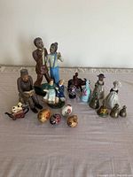 Collection of 15 figurines and decorative items arrayed on a beige surface with visible details of painted ceramic and metal bells.