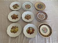 Nine decorative plates laid out on table, showing various fruit and floral designs with scalloped edges.