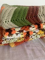 Close-up photo showing crocheted blankets stacked, with multicolor patterns in green, brown, orange, yellow, and neutral hues