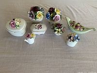 Top view of 7 small intricate bone china figurines with spring flowers including roses and pansies in various colors. Items include small pots, leaf dish, watering can, and boxes.