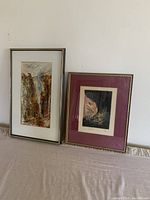 Two framed prints displayed side-by-side, one with a white mat and abstract earth-tone image, the other with a red mat and dark abstract image in gold-tone metal frames.